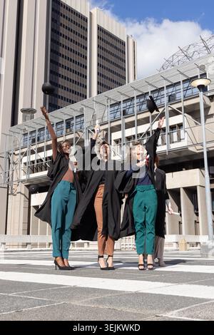 Diverse female graduates in black gowns tossing caps, holding diploma on patterned plaza by tower. Mortarboard, scroll, urban, c Stock Photo