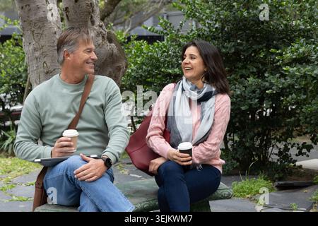 Middle-aged couple sitting on stone bench in urban green space, holding takeaway cups and tablet. Adult, companions, conversatio Stock Photo
