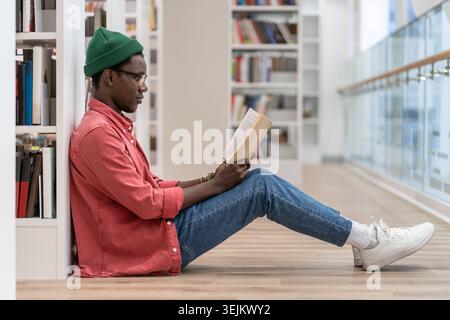 Focused Black student man book lover spending free leisure time in library, African American guy with book in hands sits on floor near bookcase enjoyi Stock Photo