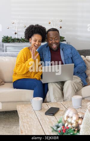 African American couple sitting on sofa at home, man in denim jacket holding laptop, woman waving. Living, room, cozy, festive, rustic, decor, christm Stock Photo