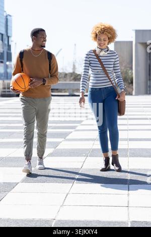African American friends walking across plaza carrying basketball, wearing backpacks and headphones. Urban, sunny, casual, pedes Stock Photo