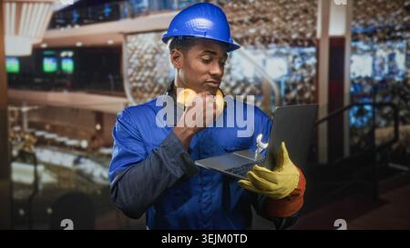 Man in hardhat wearing blue coverall and noise cancel headphones holding laptop with wrench and hand to chin in building; focused problem solving. Stock Photo