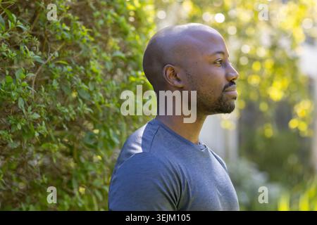 Adult African American man standing in sunlit yard by hedge with bokeh, wearing tee, copy space Stock Photo