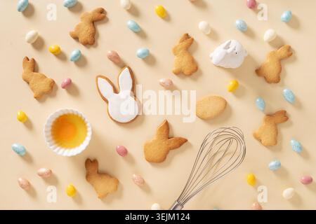 Easter pattern with bunnies shaped cookies, eggs, sweet chocolate eggs, themed cookie cutters and whisk on beige background. Festive food and snacks. Stock Photo