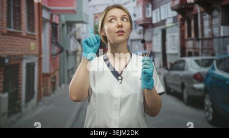 Woman dentist in uniform holding dental mirror and probes, pointing index finger while wearing blue gloves on street; reassurance care trust. Stock Photo
