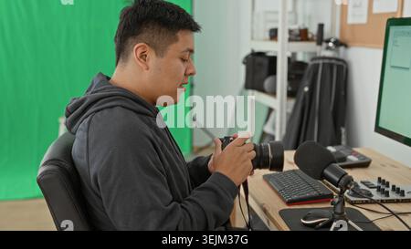 Man sits at desk adjusting camera lens in studio with green screen, keyboard, microphone and monitor visible; quiet calm. Stock Photo