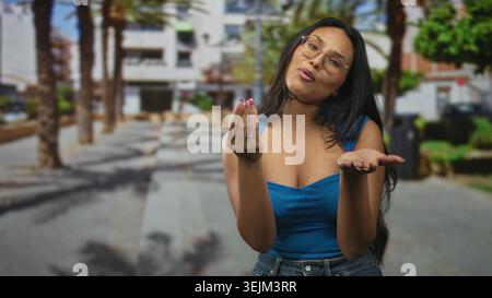 Woman in blue top beckons with both hands and open palms while tilting her head in a palm tree lined street; questioning curiosity. Stock Photo
