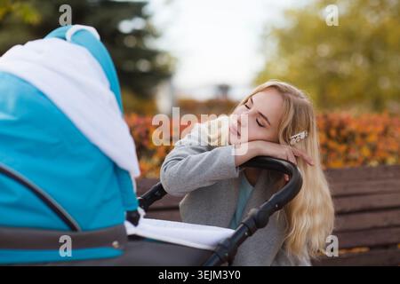 Depression young woman with baby carriage sleeps sitting on a bench in the park. frustrated mum tired of baby Stock Photo
