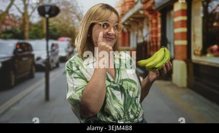 Middle age hispanic blonde woman holds bananas in one hand and points finger to glasses with the other on a street with parked cars and storefronts; p Stock Photo