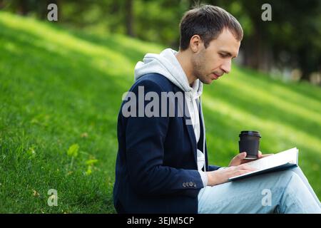 Casual businessman read book and drink coffee outdoors sitting on green grass in park during break Stock Photo