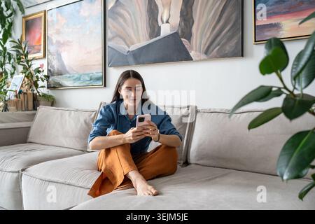 Curious woman artist sitting on couch watching media online on smartphone at cozy home gallery. Stock Photo