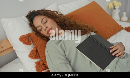 Woman sleeping in a bedroom, hand resting on a closed black notebook on her chest, eyes closed and curly hair spread on pillow; quiet calm. Stock Photo