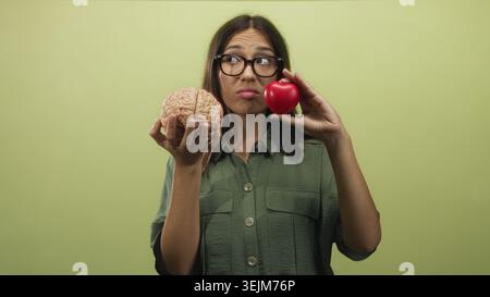 Young hispanic brunette woman holding a brain model and red heart stress ball in hands against green studio wall; indecision. Stock Photo
