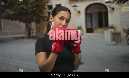 Young woman with red boxing gloves holding guard pose on a street in front of a stone building; determination grit. Stock Photo