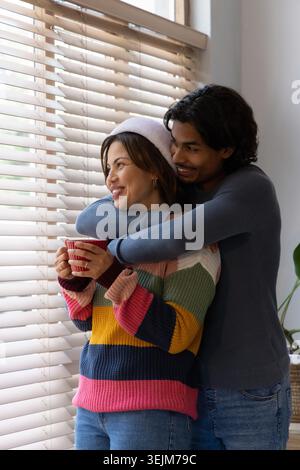 Couple embracing while woman holding striped mug in striped sweater and beanie at home by blinds. Knitwear, romance, hug, cup, w Stock Photo