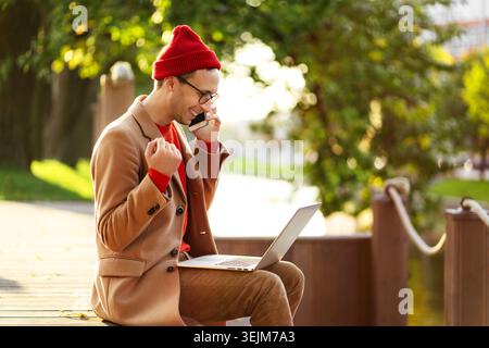 Smiling freelancer man wear glasses talking on mobile phone, sitting outdoors, working on laptop Stock Photo