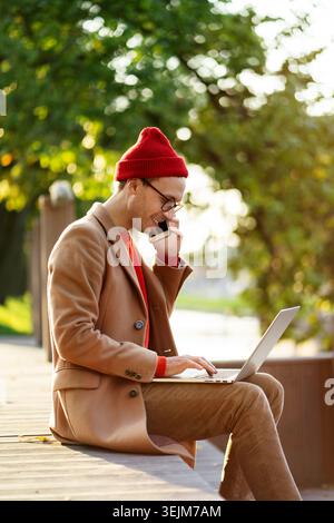 Smiling freelancer man wear glasses talking on mobile phone, working on laptop, sitting outdoors. Stock Photo