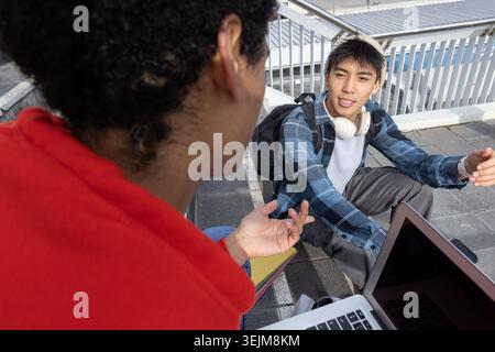 Diverse male students discussing class notes on concrete steps at campus with laptop and headphones. Discussion, study, collabor Stock Photo