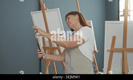 Woman painter selects paintbrushes and adjusts brush tips before blank canvas in studio; creative focus. Stock Photo