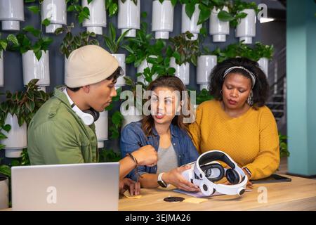 Diverse coworkers discussing VR headset demo at wooden communal table with laptop and notes. Collaboration, innovation, technology, coworking, greener Stock Photo
