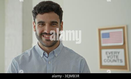 Man smiling with visible face and beard, looking at camera in a building with an american flag poster and 'every vote counts' sign; civic duty hopeful Stock Photo