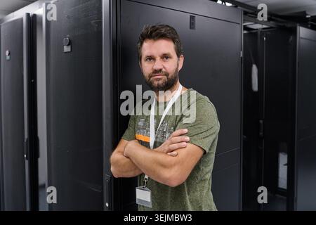 A confident data center technician stands with arms crossed in a server room beside black server racks, wearing an ID badge; conveys professionalism, Stock Photo