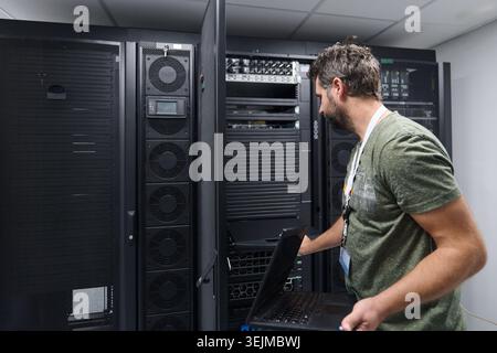 IT technician working on server rack in data center with laptop performing network maintenance Stock Photo