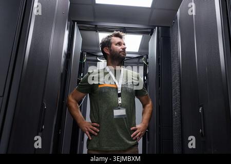 Male IT technician wearing an ID badge stands confidently between server racks in a modern data center, inspecting equipment and monitoring network in Stock Photo