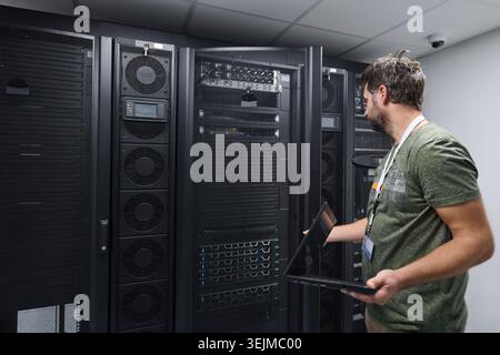 IT technician inspecting servers in a data center with a laptop, performing maintenance and network checks Stock Photo