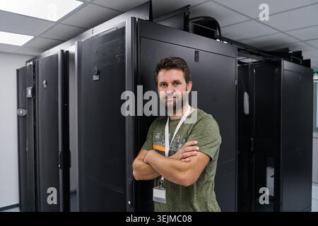 IT technician standing confidently in a data center next to server racks wearing an ID badge Stock Photo