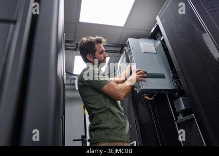 A focused data center technician installs a rack-mount server and connects power and network cables in a secure server room, performing routine hardwa Stock Photo