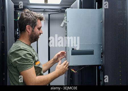 An IT technician inspects and installs server hardware inside a data center rack, checking cables and connections during routine maintenance to ensure Stock Photo