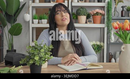 Woman florist in apron smiling with hands clasped on open notebook at studio counter, surrounded by potted plants, vases, shelves and tulips; welcomin Stock Photo