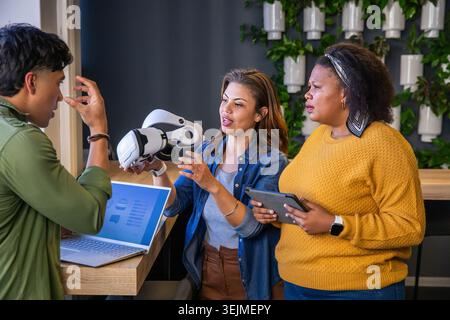 Diverse coworkers demonstrating white VR headset at wooden counter with open laptop and tablet. Collaboration, innovation, techn Stock Photo