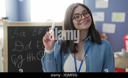 Young hispanic woman holding chalk and smiling in a classroom with a blackboard, alphabet letters, posters and a lanyard badge; joyful learning. Stock Photo