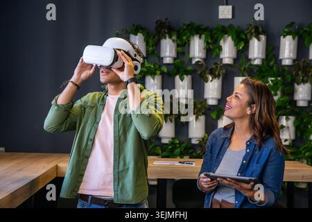 Diverse coworkers adjusting white VR headset and holding tablet at modern office wooden high table. Coworking, teamwork, virtual Stock Photo
