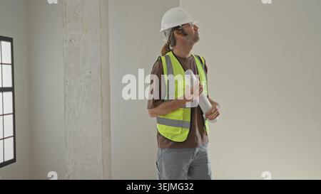Man in hardhat and reflective vest holding rolled blueprint and looking up inside building; planning concentration. Stock Photo