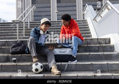 Diverse male students sitting on campus steps, studying tablet, one wearing red hoodie. Urban, plaza, staircase, teamwork, study Stock Photo
