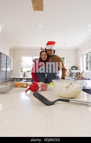 Romantic couple wearing striped aprons and festive hats baking at kitchen island with mixing bowl. Cozy, warm, cooking, home, interior, sunlit, togeth Stock Photo