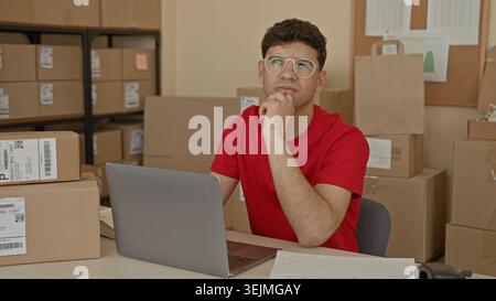 Man in red shirt typing on laptop amid stacked cardboard packages at a packing workstation in building; entrepreneurship concentration. Stock Photo