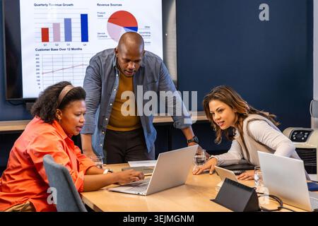 Diverse coworkers in business casual discussing charts in conference room using laptops and tablet. Boardroom, collaboration, focus, data, analytics, Stock Photo