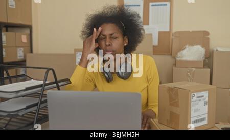 Woman with laptop, stacked parcels and headphones, hand on temple while sorting boxes at a packing building; stress overwhelm. Stock Photo