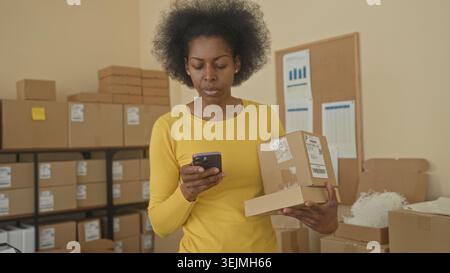 Woman holding parcel and smartphone, hand tapping screen while checking shipping labels in a warehouse packing room; entrepreneurial focus. Stock Photo