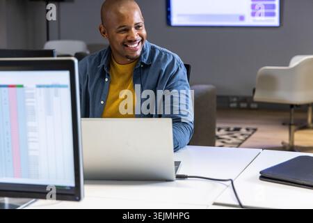 African American man using laptop at office smiling, glancing at spreadsheet monitor in mustard top. Coworking, productive, mode Stock Photo