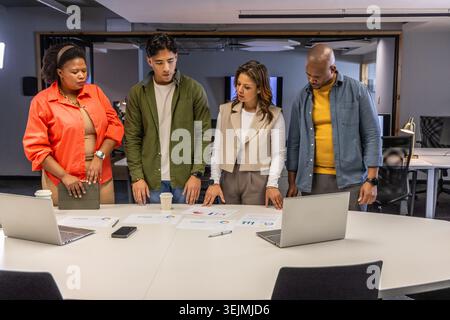 Diverse coworkers in business clothes reviewing charts and two laptops at modern office table. Collaboration, monitors, tablet, Stock Photo