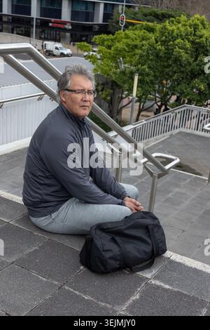 Senior Asian man sitting on concrete steps wearing glasses by metal handrail with black duffel bag. Urban, solitary, contemplative, overcast, pedestri Stock Photo
