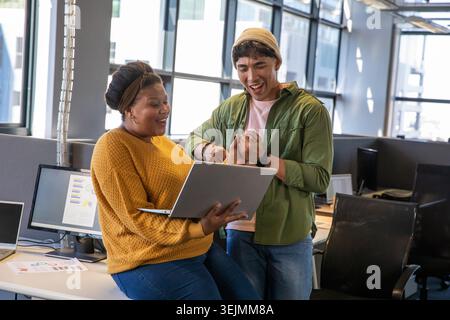 Diverse coworkers standing at workstation, leaning in and celebrating over silver laptop and charts. Collaboration, teamwork, of Stock Photo