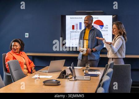 Diverse team in business wear presenting and reacting at meeting table with wall screen and laptops. Boardroom, collaboration, p Stock Photo