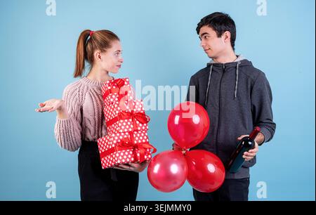 Puzzled young couple holding a pile of gift boxes, balloons and a bottle of wine looking at each other on blue background Stock Photo