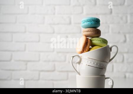 Stack of white coffee cups filled with macaroons on the edge of the table, white brick wall background Stock Photo
